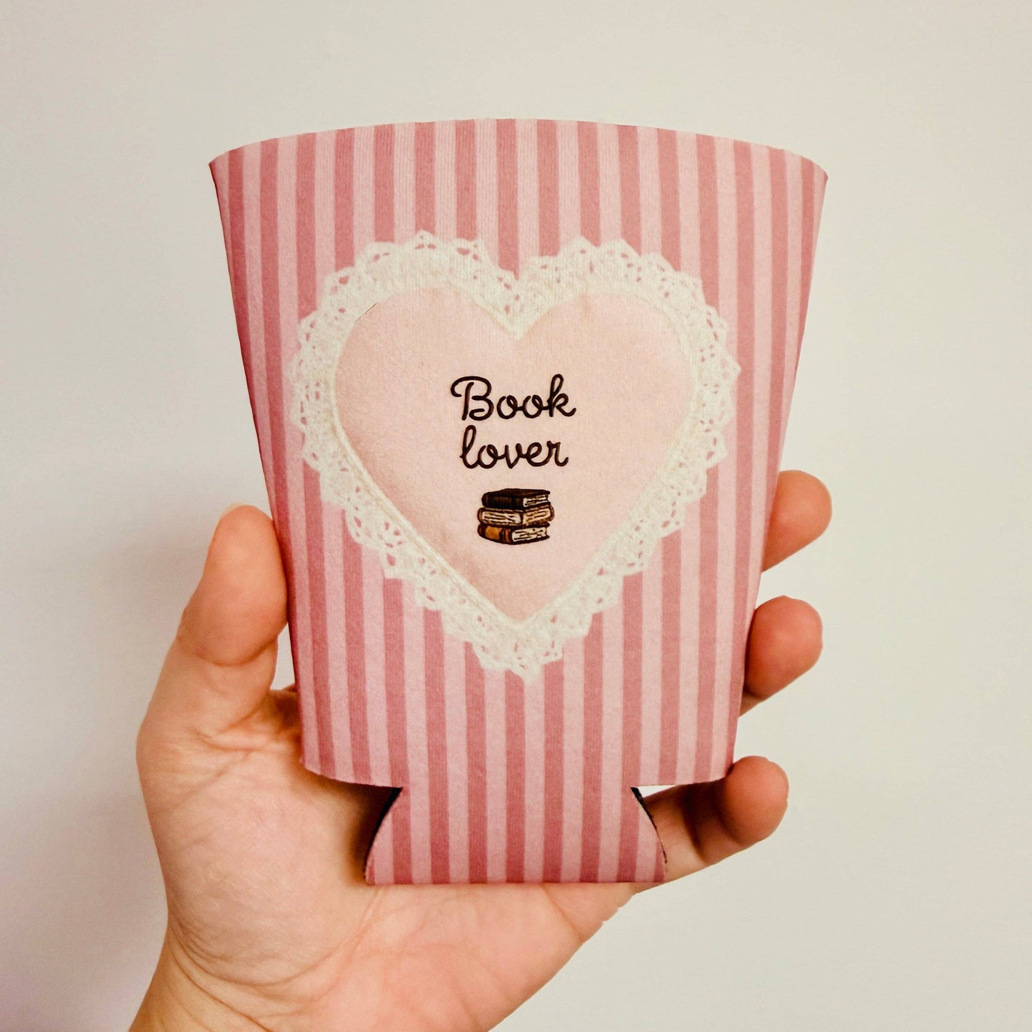 Pink striped can cooler with heart design and 'Book Lover' text held in a hand.