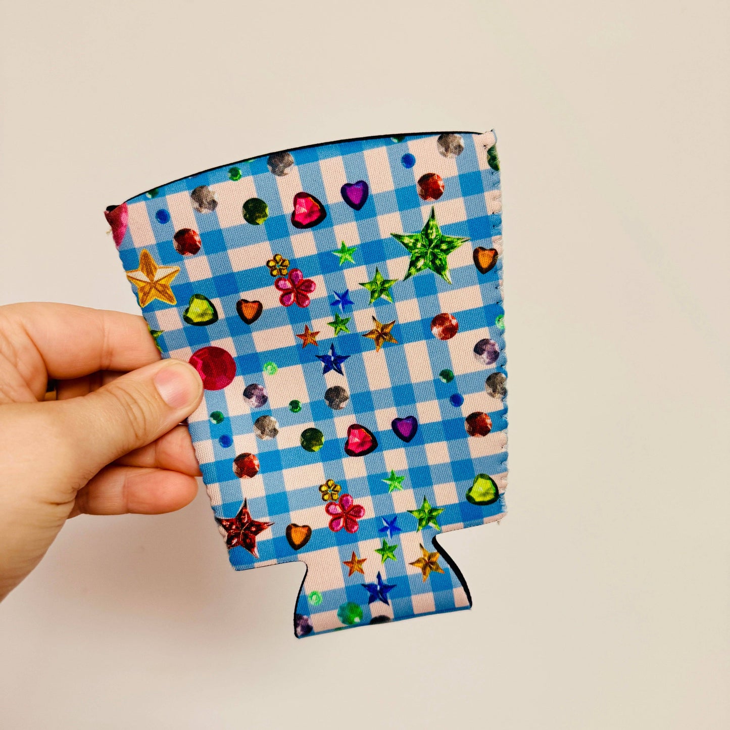 Hand holding a colorful can cooler with a blue checkered pattern and star, heart, and flower designs.