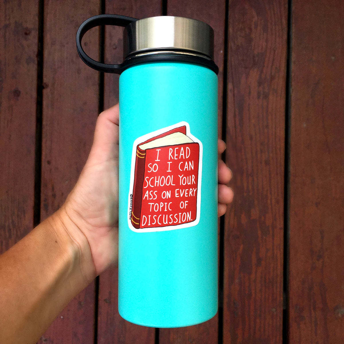 A hand holding a turquoise insulated water bottle with a stainless steel lid and black handle, featuring a red book sticker with the text 'I READ SO I CAN SCHOOL YOUR ASS ON EVERY TOPIC OF DISCUSSION.' The background is dark brown wooden siding.
