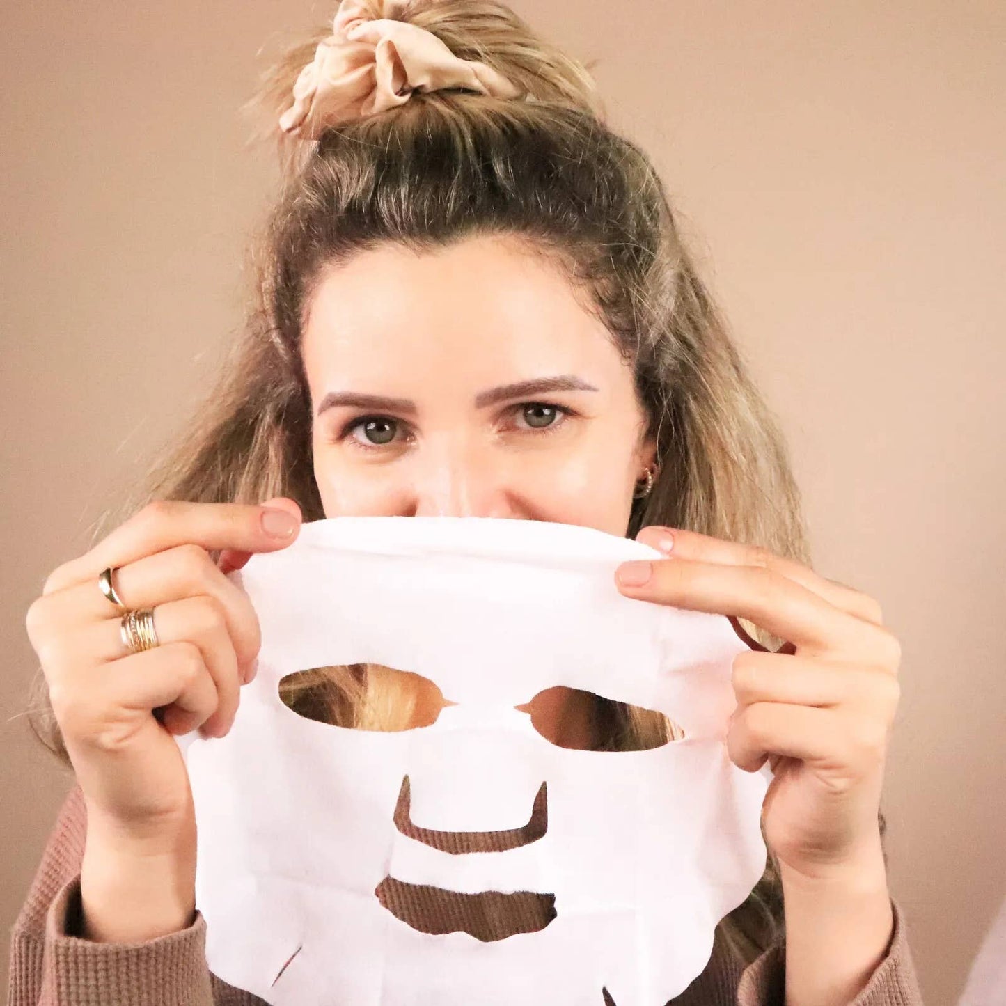 Woman holding a white face mask with cutouts over her face against a beige background