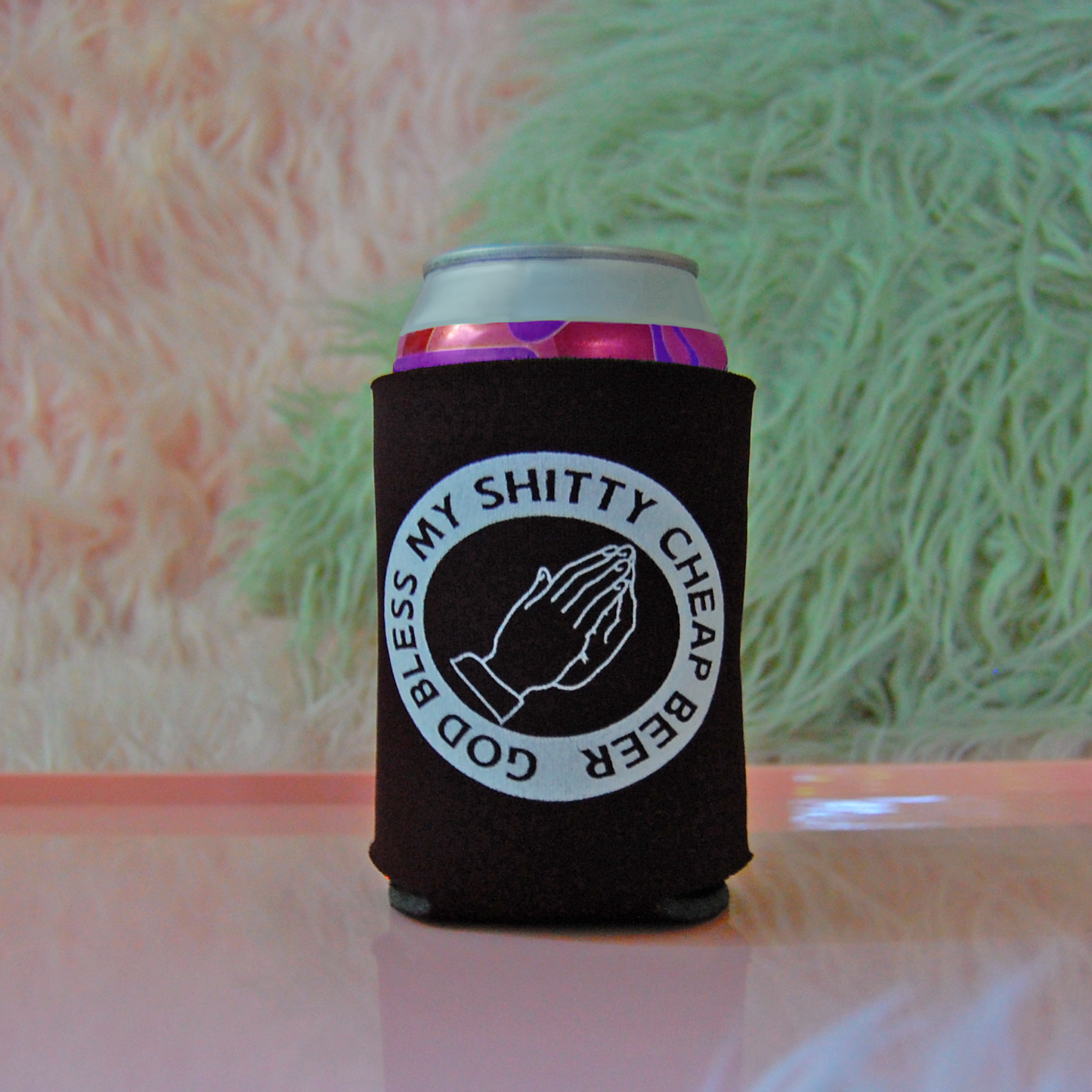 Black can cozy with a design of two praying hands and text saying "God Bless My Shitty Cheap Beer" held against a pink and green background
