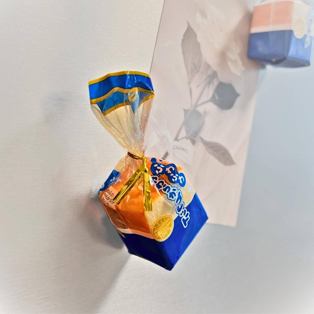 A small wrapped candy or snack package attached to a white wall, with a translucent wrapper in blue, yellow, and orange and a blue-and-white box below it. A floral card and part of another object are visible in the background.