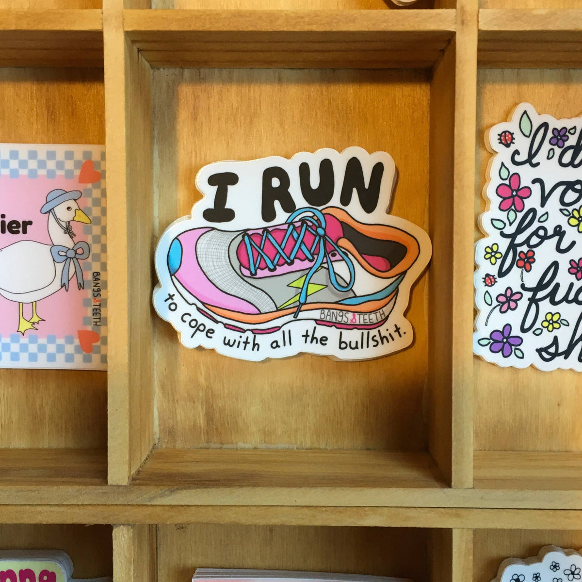 A die-cut sticker featuring a colorful running shoe with the text 'I RUN to cope with all the bullshit.' displayed on a wooden shelf alongside other stickers.