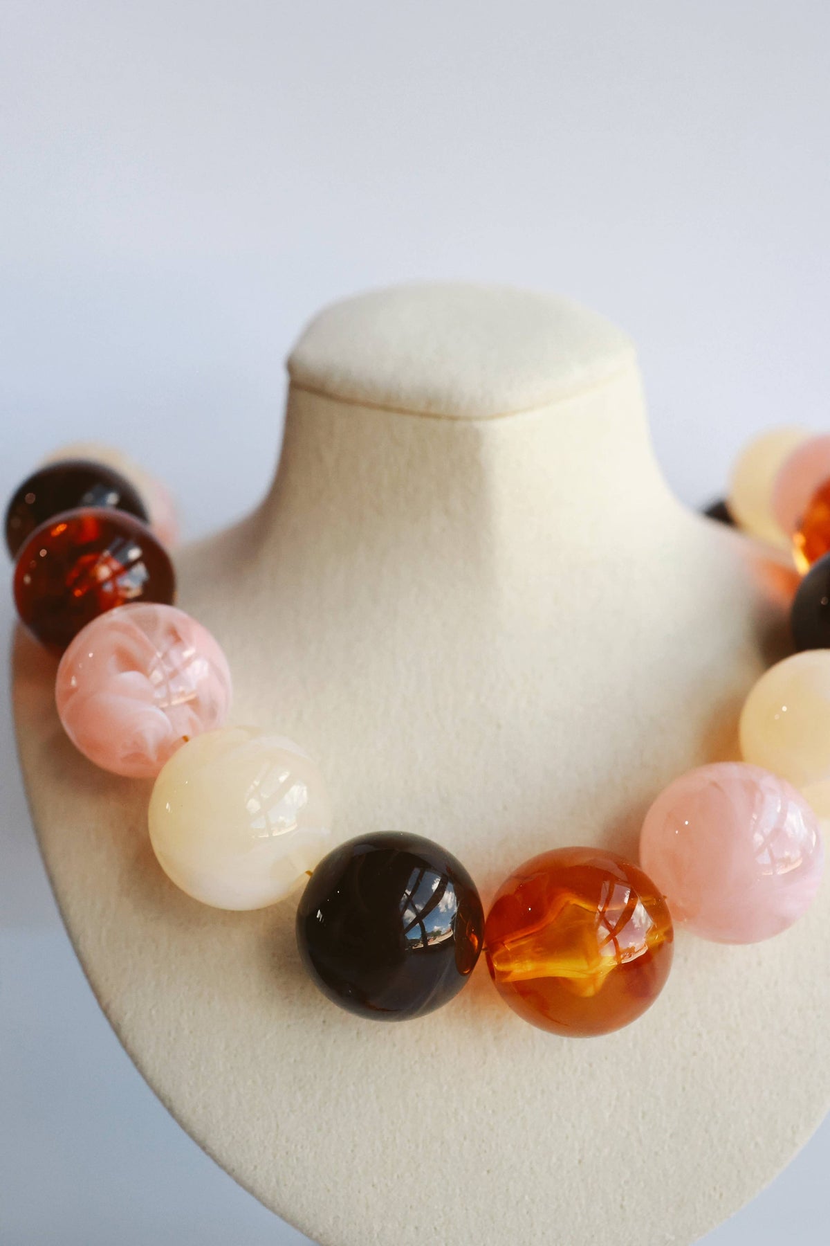 A beaded statement necklace with large, polished round beads in shades of amber orange, pale pink, cream, and dark brown, displayed on a cream-colored jewelry bust.