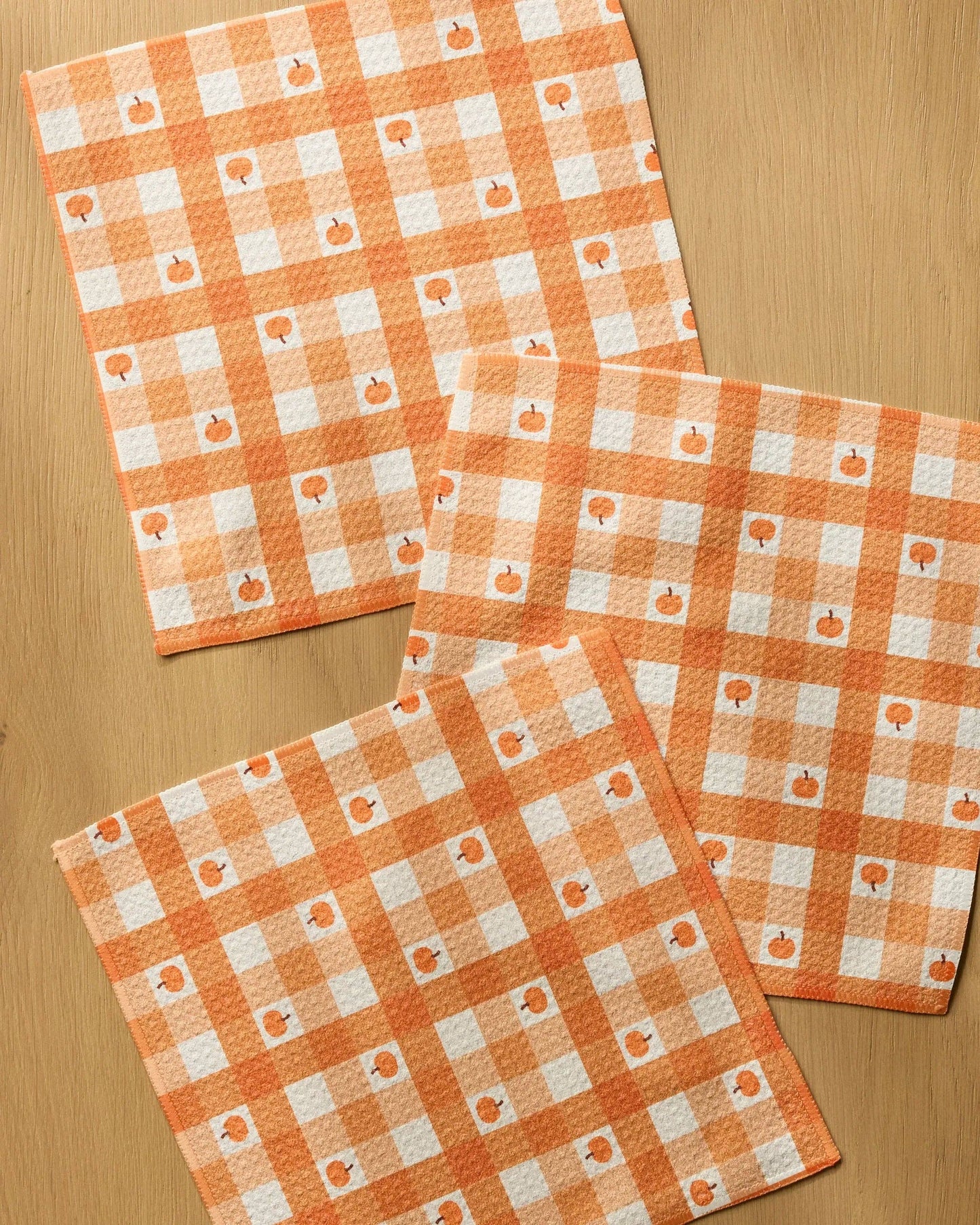 Three square kitchen towels with an orange and white gingham pattern and small pumpkin motifs, arranged on a light wood surface.