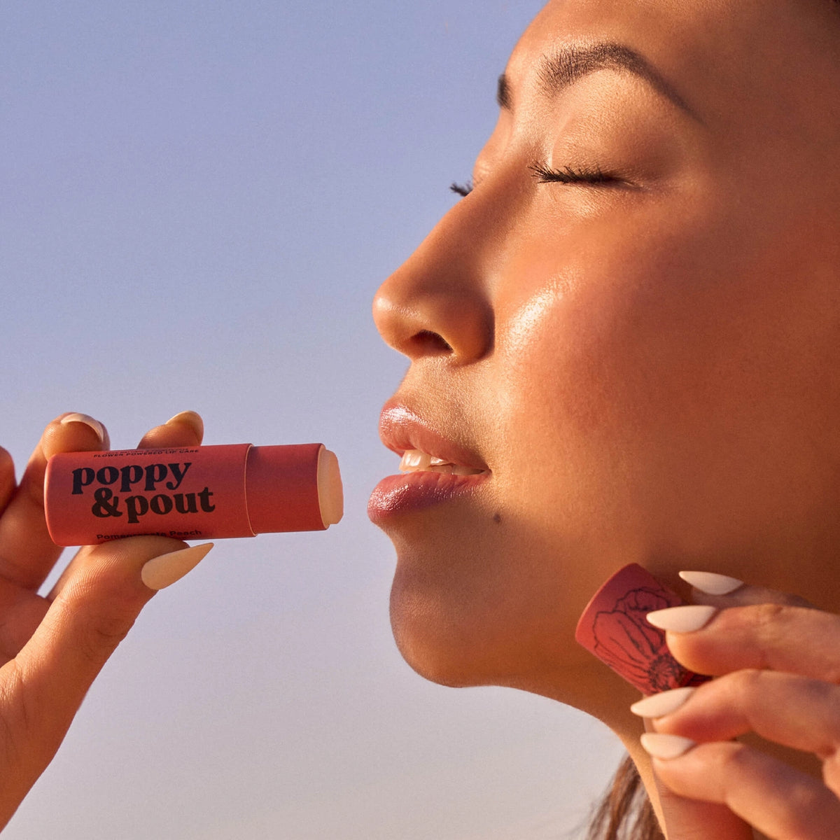 Woman applying poppy & pout lip balm against a clear blue sky