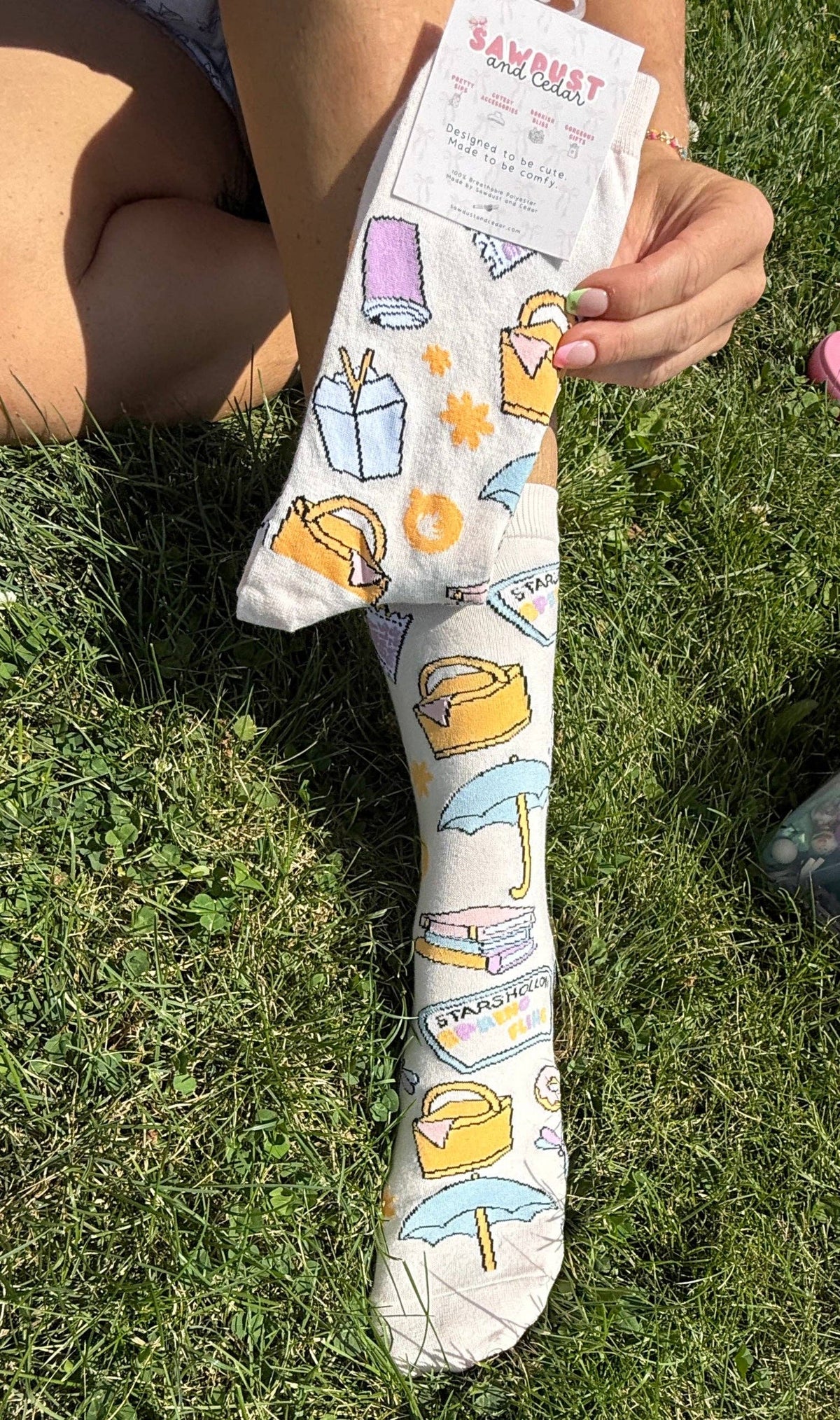 A person sitting on grass holds up a white sock with a colorful illustrated pattern of shopping bags, umbrellas, and gift boxes. The sock has a paper tag attached at the top.
