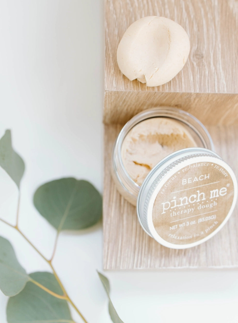 Jar of 'pinch me' therapy dough with a scoop on a wooden board, surrounded by eucalyptus leaves.