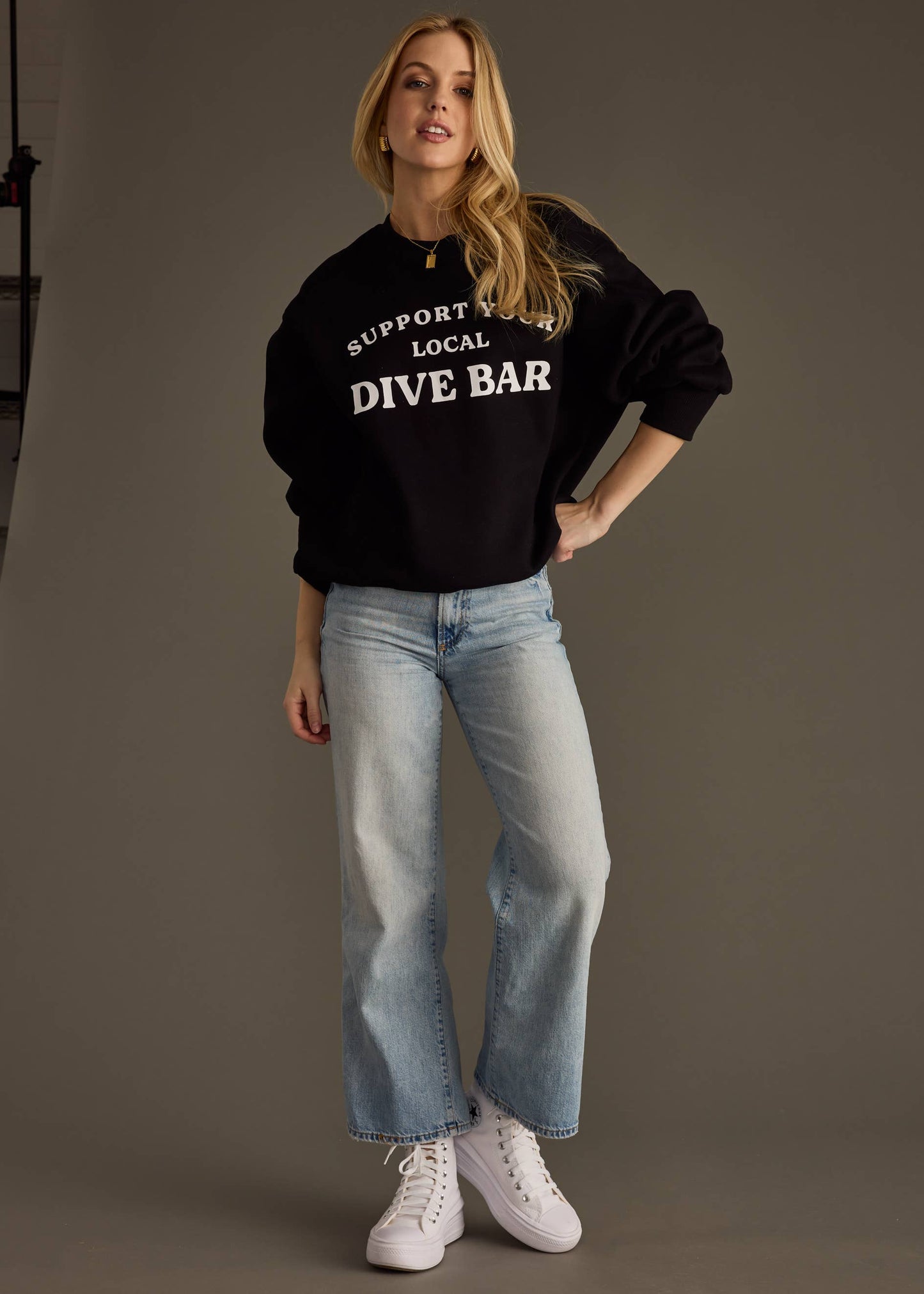Woman wearing a black sweatshirt with 'Support Local Dive Bar' text, light blue jeans, and white sneakers on a plain background.
