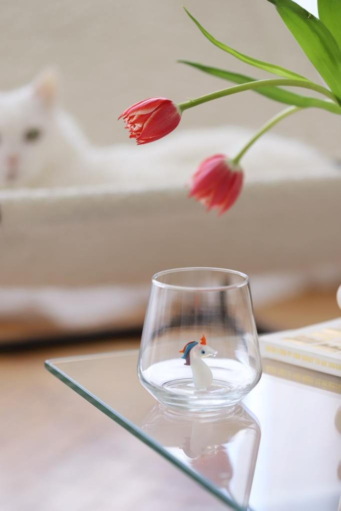 Clear glass with a small figurine of a unicorn on a reflective surface, with a blurred background.