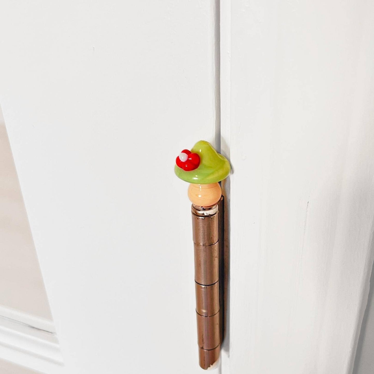 A decorative door hinge cover or door stopper in the shape of a mushroom, mounted on a brown hinge on a white door.