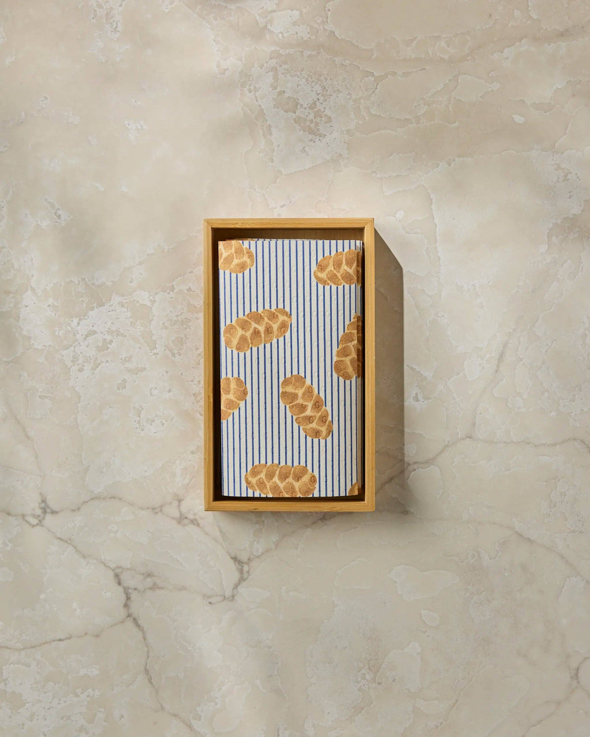 A rectangular wooden box containing a striped fabric pouch with a bread crust pattern, displayed on a textured beige marble surface.