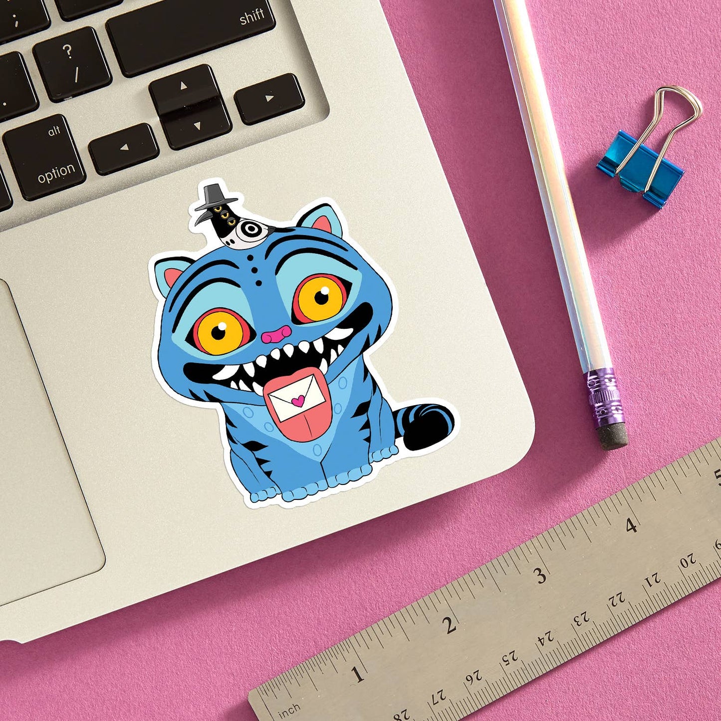 Blue cartoon cat sticker on a laptop with a pink background, ruler, pencil, and clip.