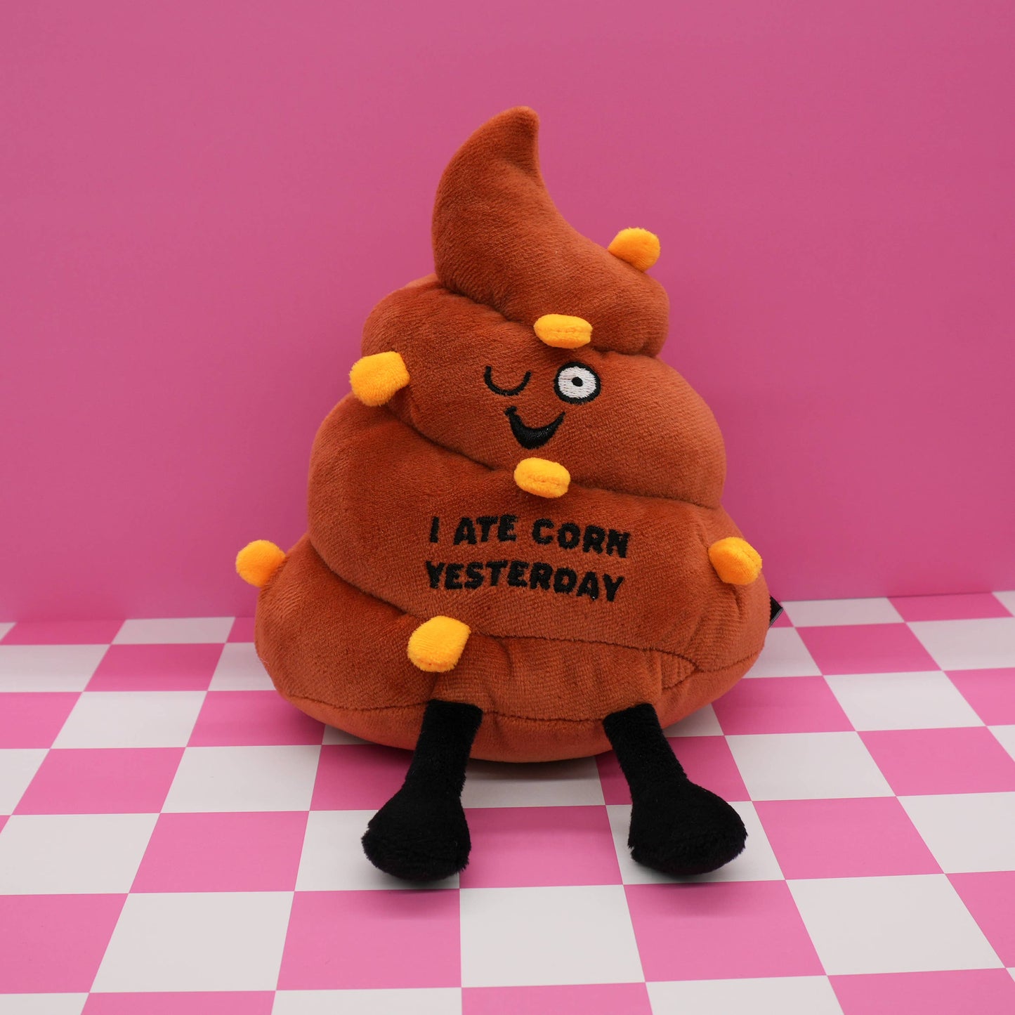 Plush toy shaped like a pile of brown poop with a face and text on a pink and white checkered floor.