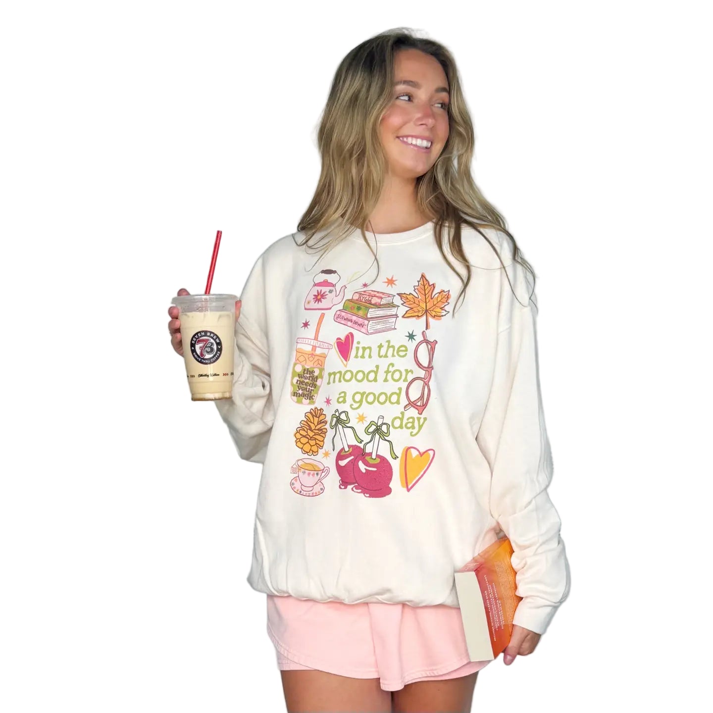 Woman wearing a white sweatshirt with colorful graphics and text, holding a 7-Brew cup and a book.