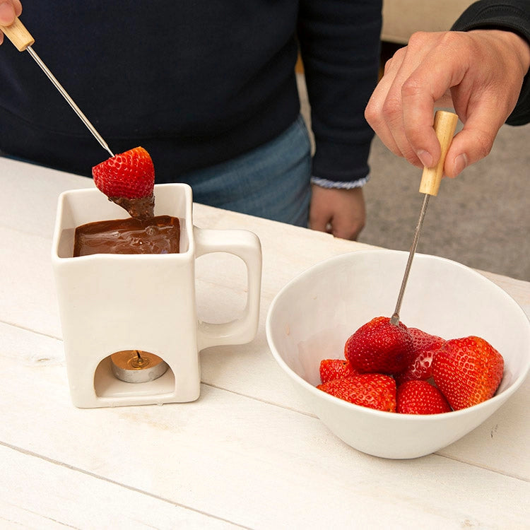 Person dipping strawberries into melted chocolate using a fondue set on a white table.