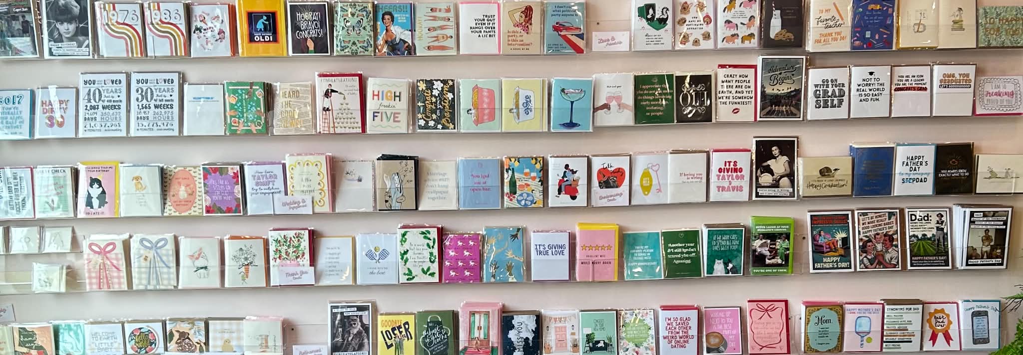 5 rows of various different greeting cards sitting on clear shelves mounted to a light pink wall. 