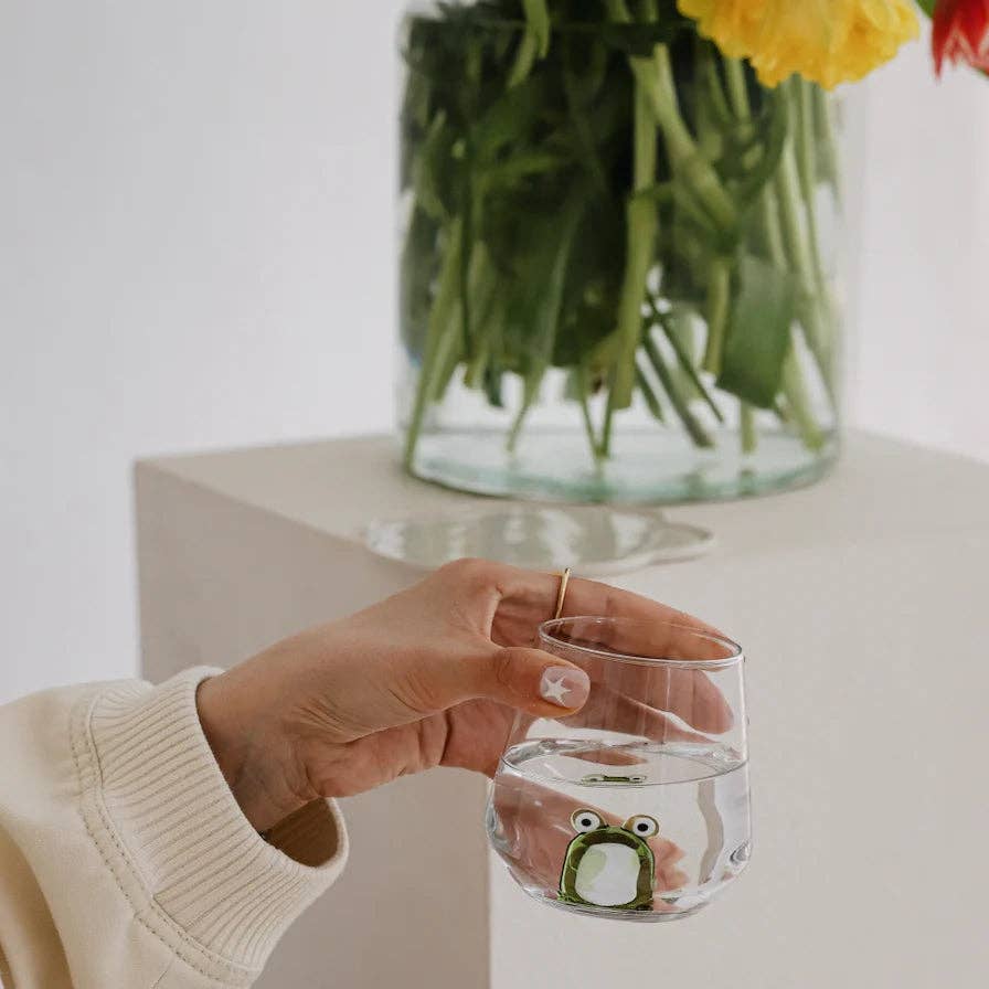 someone holding a clear glass with small frog figurine sitting in the middle with a vase of flowers behind it
