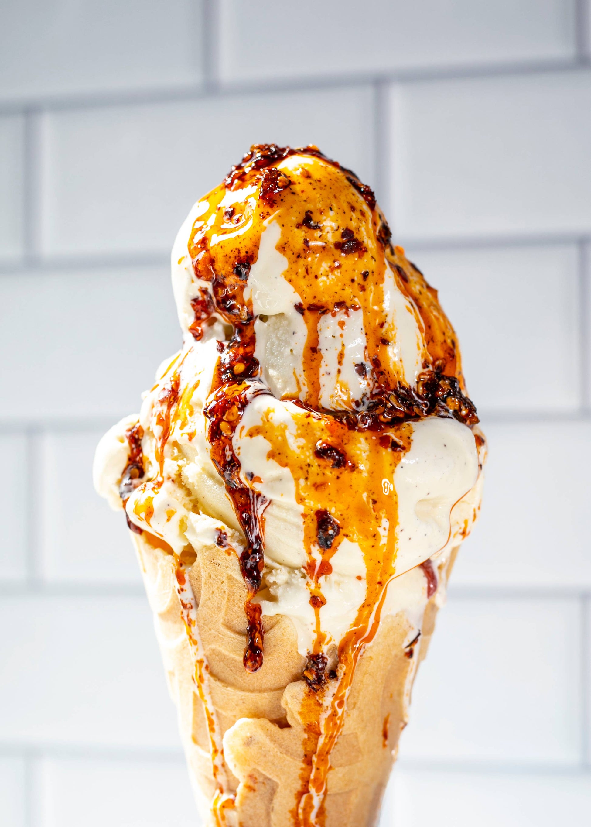 Ice cream cone with momofuku crunch drizzle against a white tiled wall.