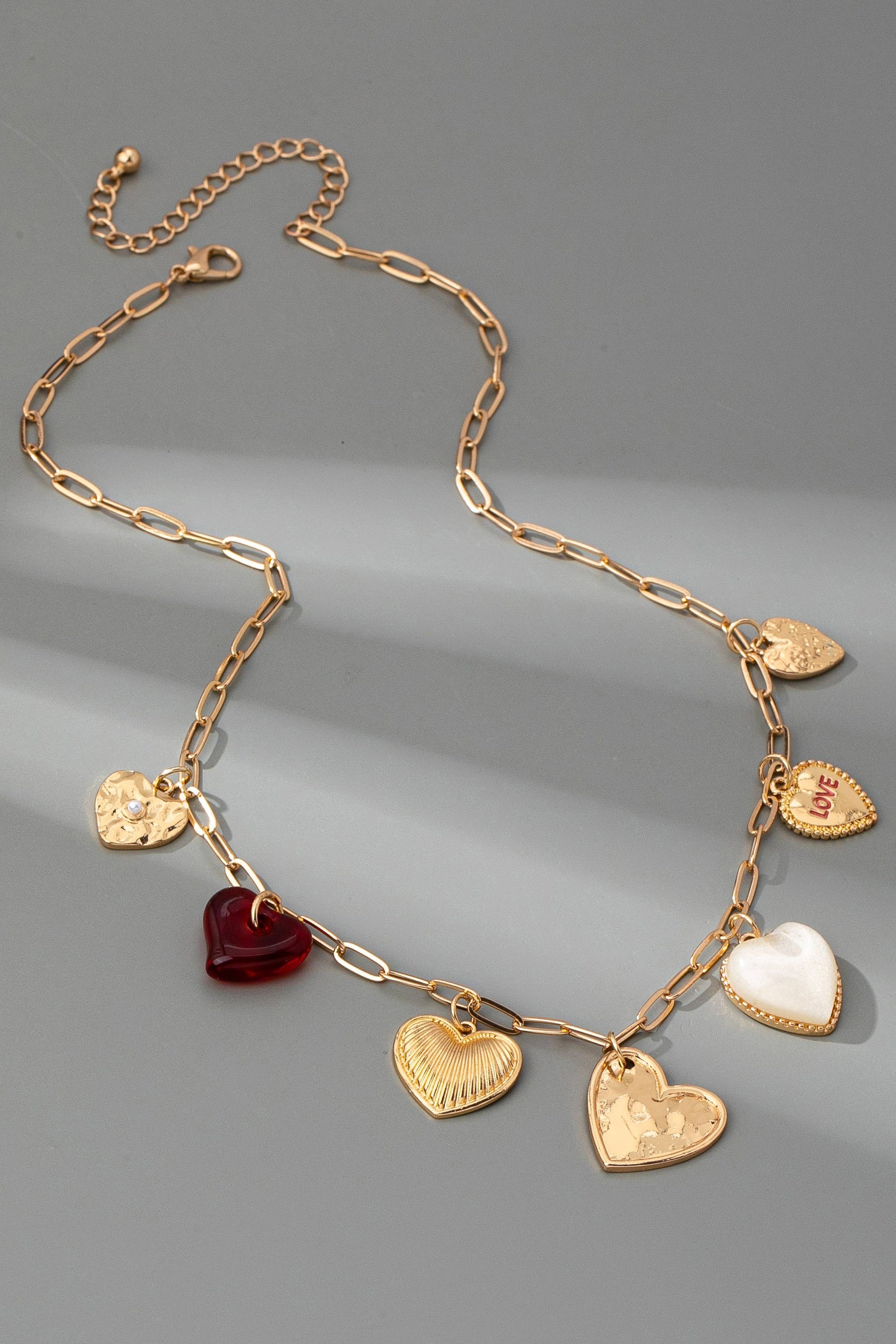 Gold chain link necklace with 5 different heart shaped gold charms, 1 pearl heart charm, and one clear red charm, on a grey background