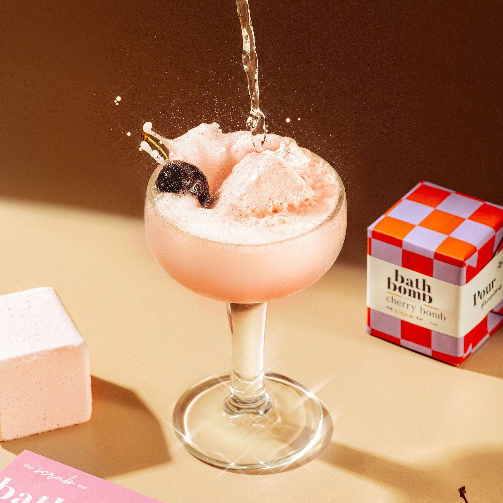 A fizzy pink bath bomb in a coupe glass with a cherry and water poured over it, with a a checkered box labeled 'bath bomb' in the background.