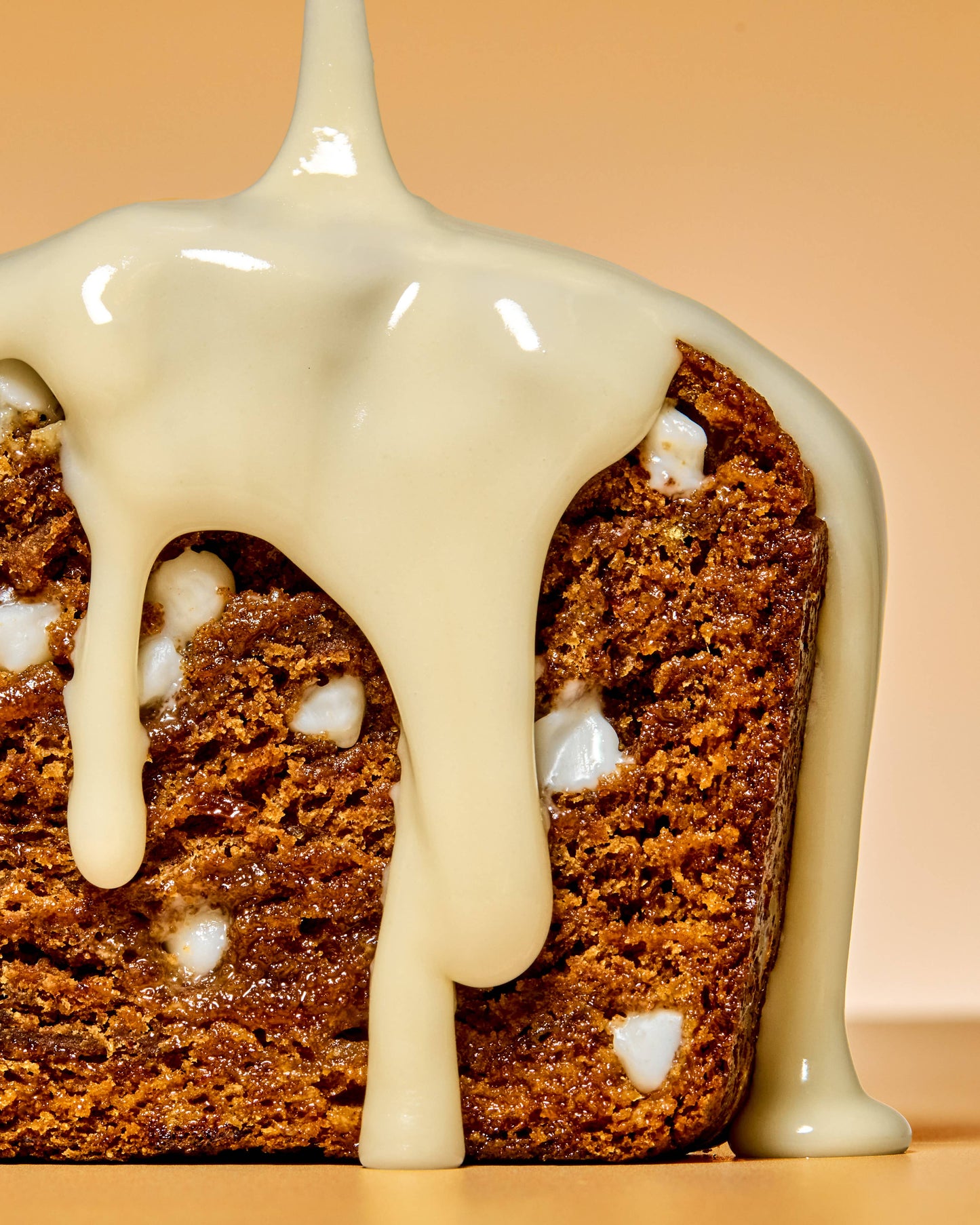 Slice of banana bread with creamy glaze being poured on it, on a peach background