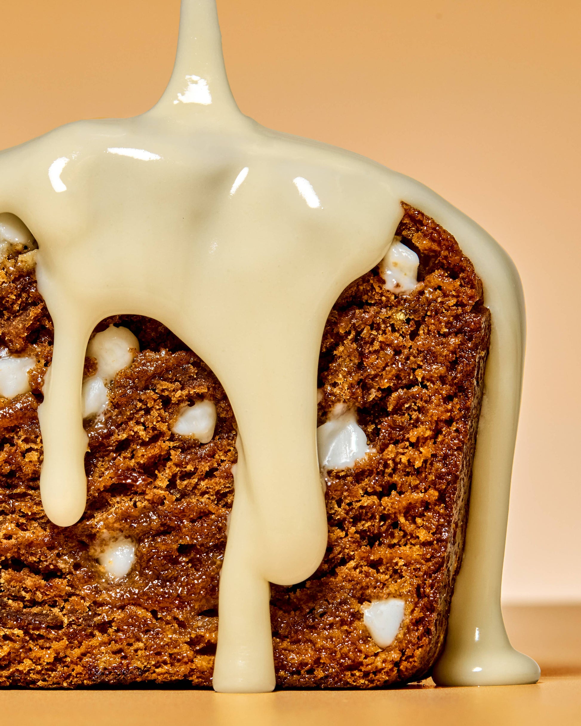 Slice of banana bread with creamy glaze being poured on it, on a peach background