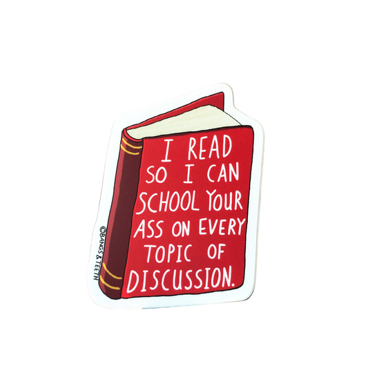 A die-cut sticker shaped like a red book with white text reading 'I READ SO I CAN SCHOOL YOUR ASS ON EVERY TOPIC OF DISCUSSION.' It is photographed standing upright on a book against a blurred brick wall background.