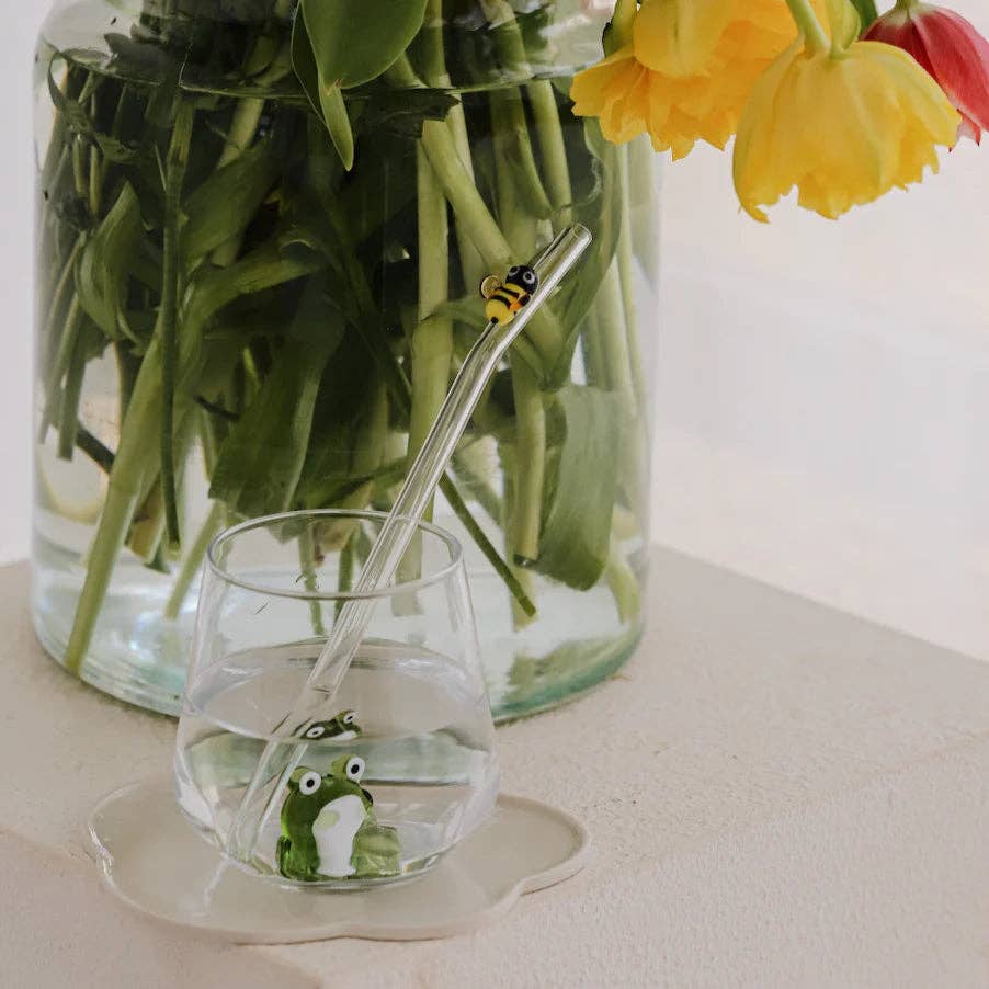 clear glass with small frog figurine sitting in the middle with a vase of flowers behind it