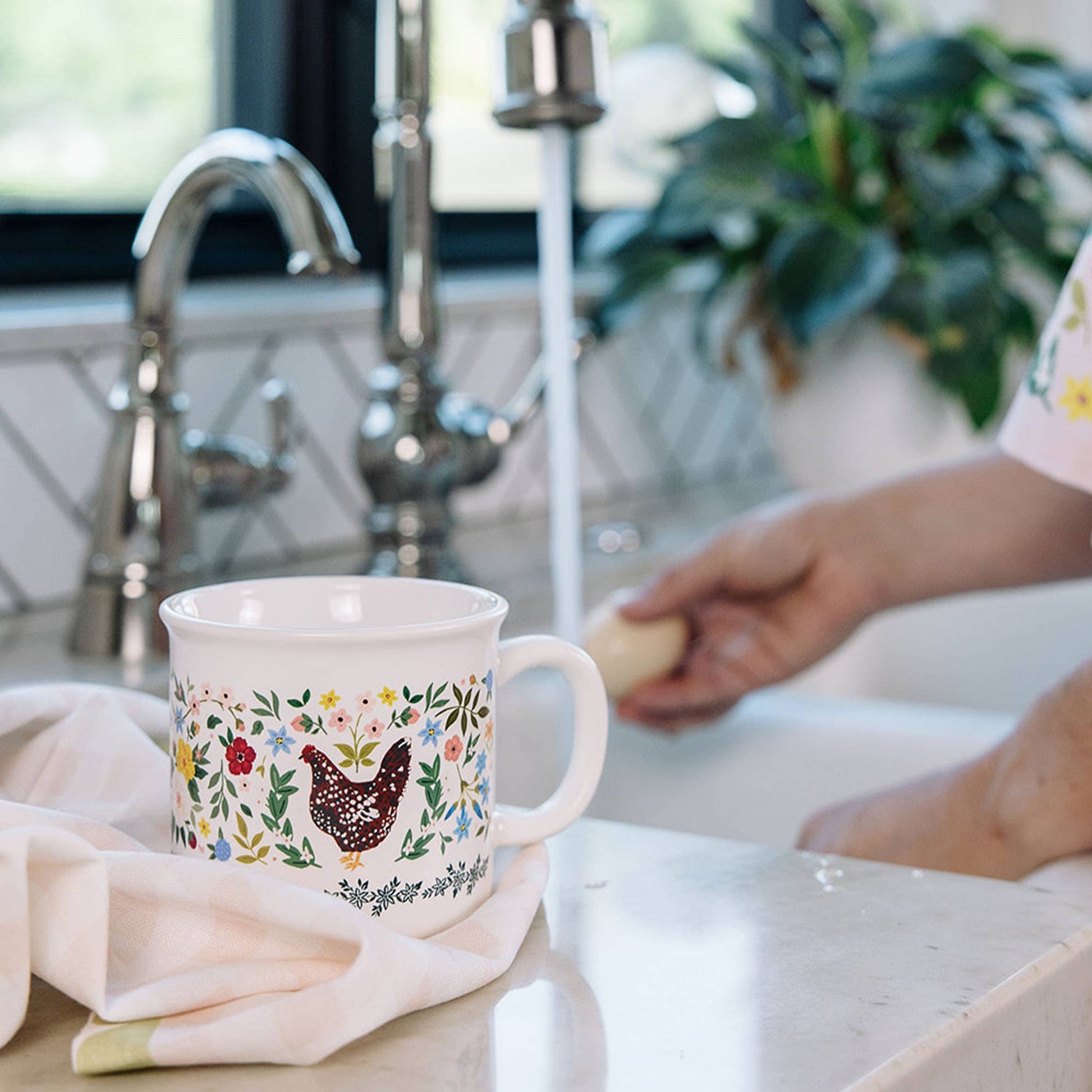 Decorative mug with floral and chicken design on a kitchen counter next to a sink.