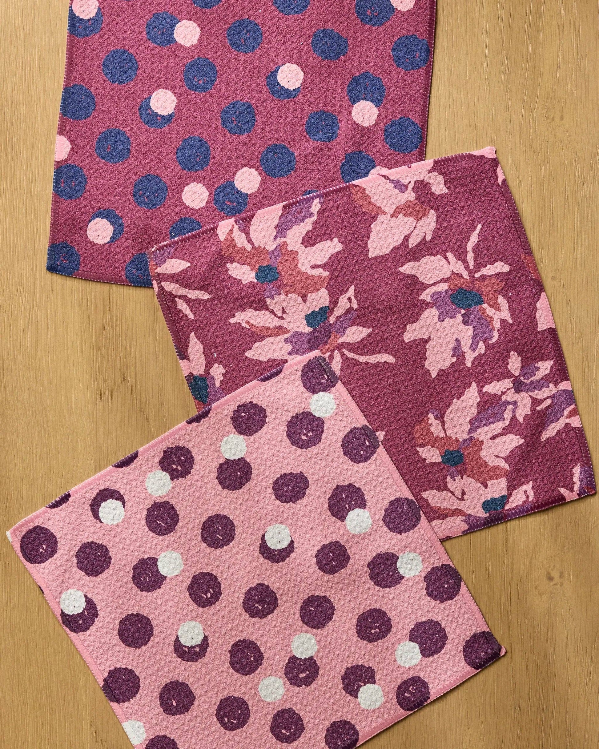 Three square fabric napkins with textured woven patterns, featuring pink, purple, and navy polka dots and floral prints, arranged on a light wood background.