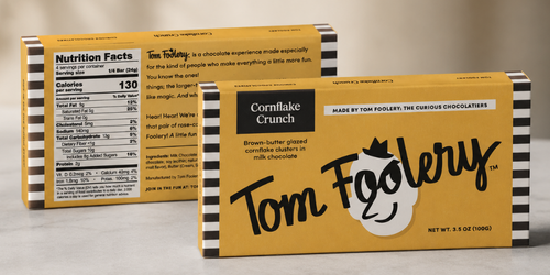 Two boxes of Tom Foolery Cornflake Crunch chocolate bars on a light gray surface, with the front box showing a yellow and black design and the back box showing the nutrition facts panel.