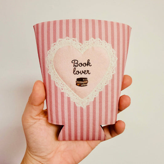 Pink striped can cooler with heart design and 'Book Lover' text held in a hand.