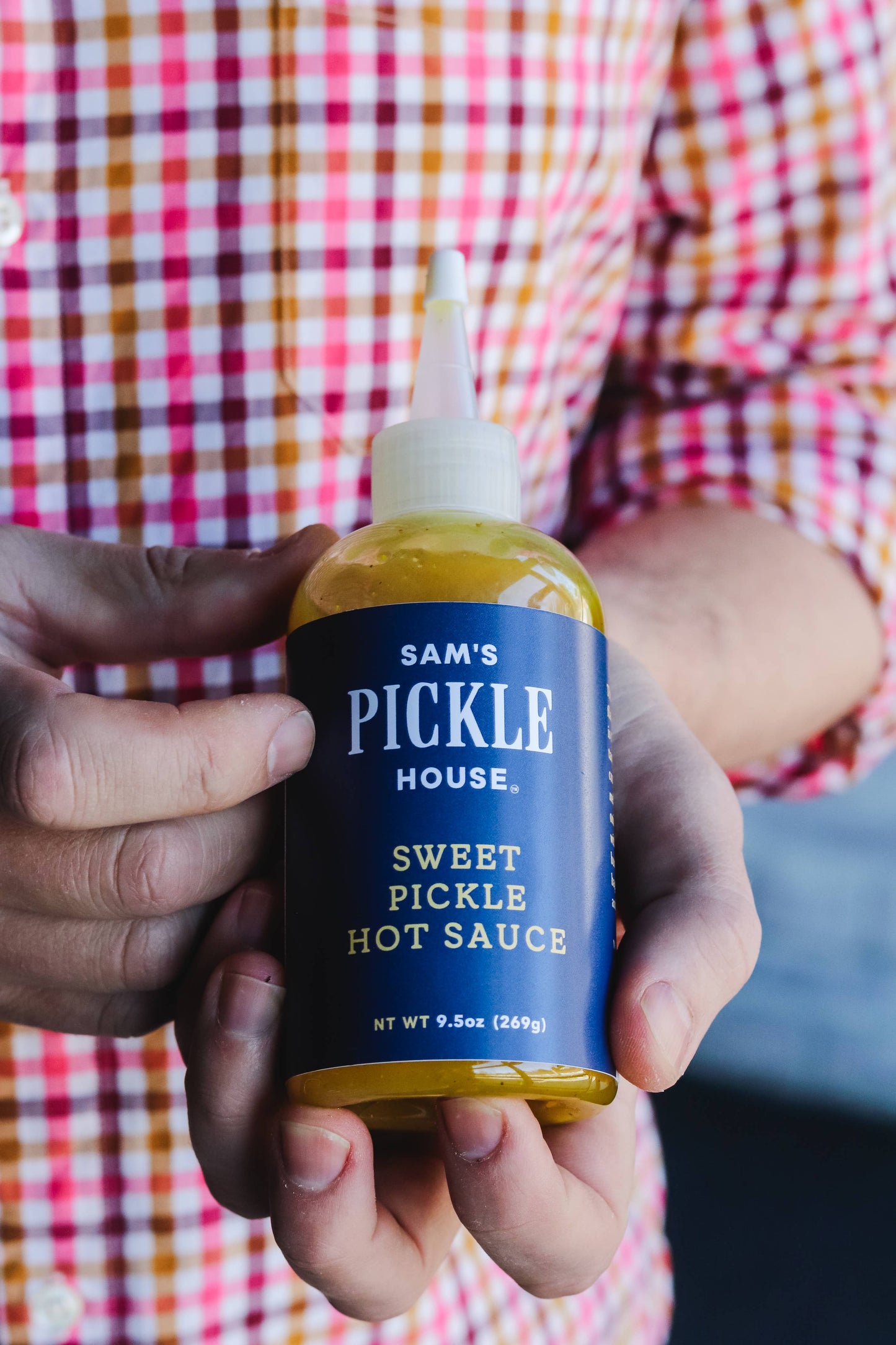 Person holding a bottle of Sam's Pickle House Sweet Pickle Hot Sauce with a checkered shirt in the background.