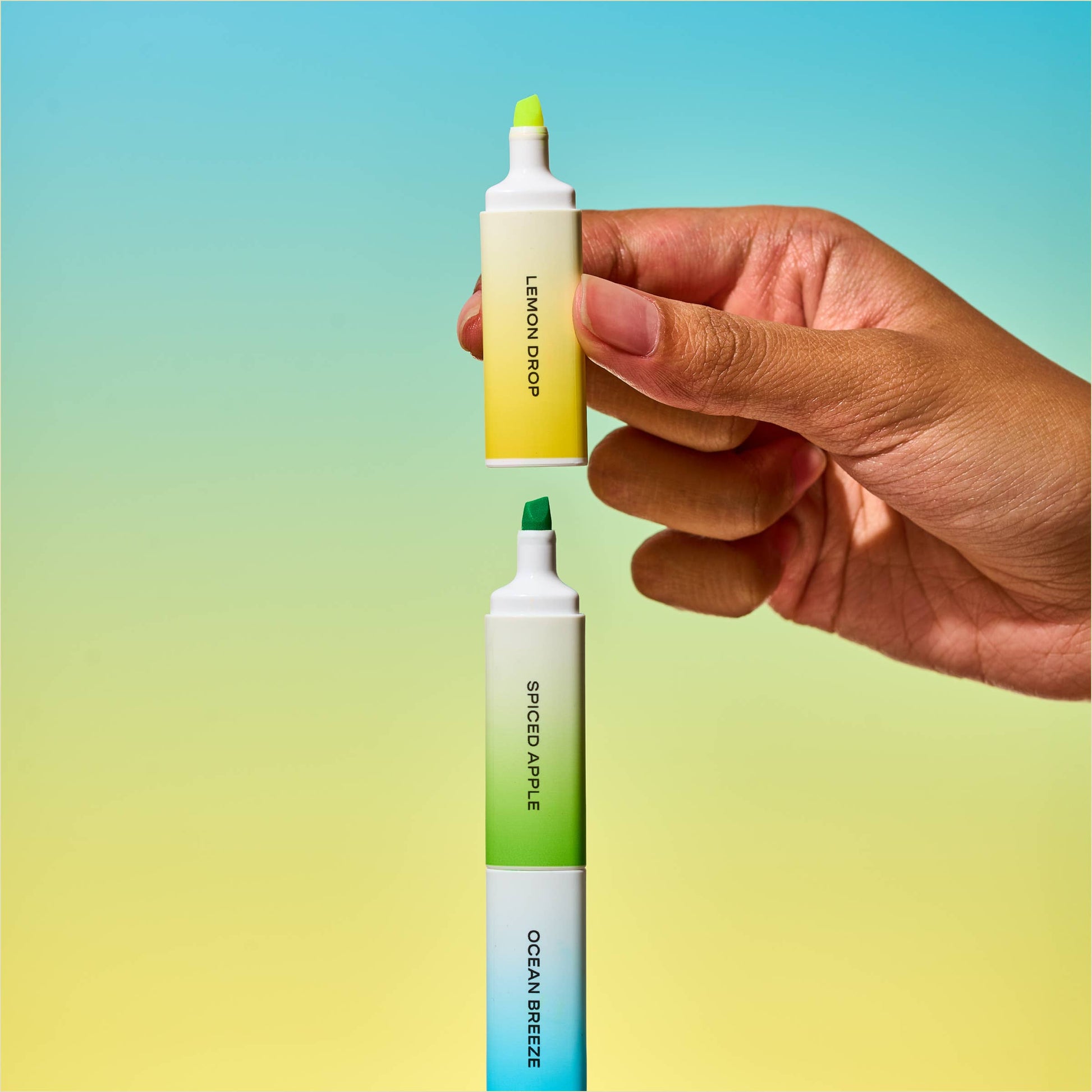 Hand holding a yellow hghlighter labeled 'lemon drop', with a green highlighter underneath labeled 'spiced apple' and a blue highlighter beneath labeled 'ocean breeze' on a blue and green gradient background