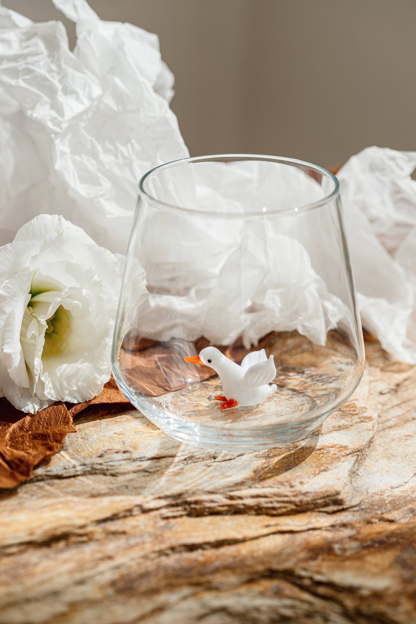 Clear glass with a small white duck figurine on a textured surface with white flowers in the background