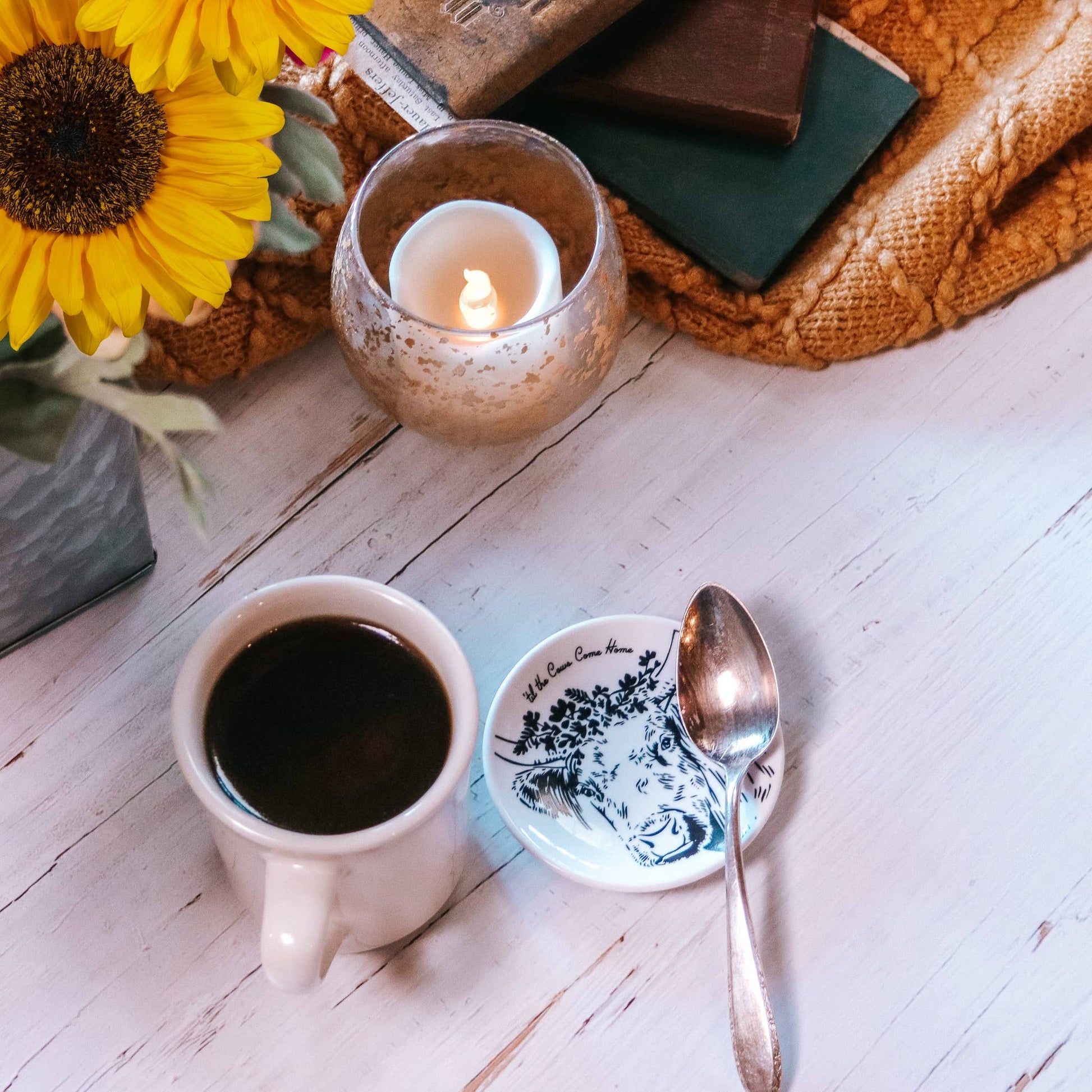 Cup of coffee, saucer with spoon, candle, and sunflowers on a wooden surface