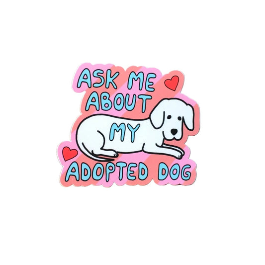 A die-cut sticker featuring a white cartoon dog and the text 'ASK ME ABOUT MY ADOPTED DOG' in pink, blue, and red, displayed on a wooden ledge against a red brick wall.