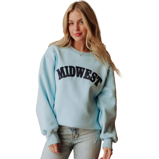 A woman wearing a light blue crewneck sweatshirt with 'MIDWEST' printed in dark navy arched letters across the chest, paired with light-wash jeans, standing in front of a beige curtain backdrop.
