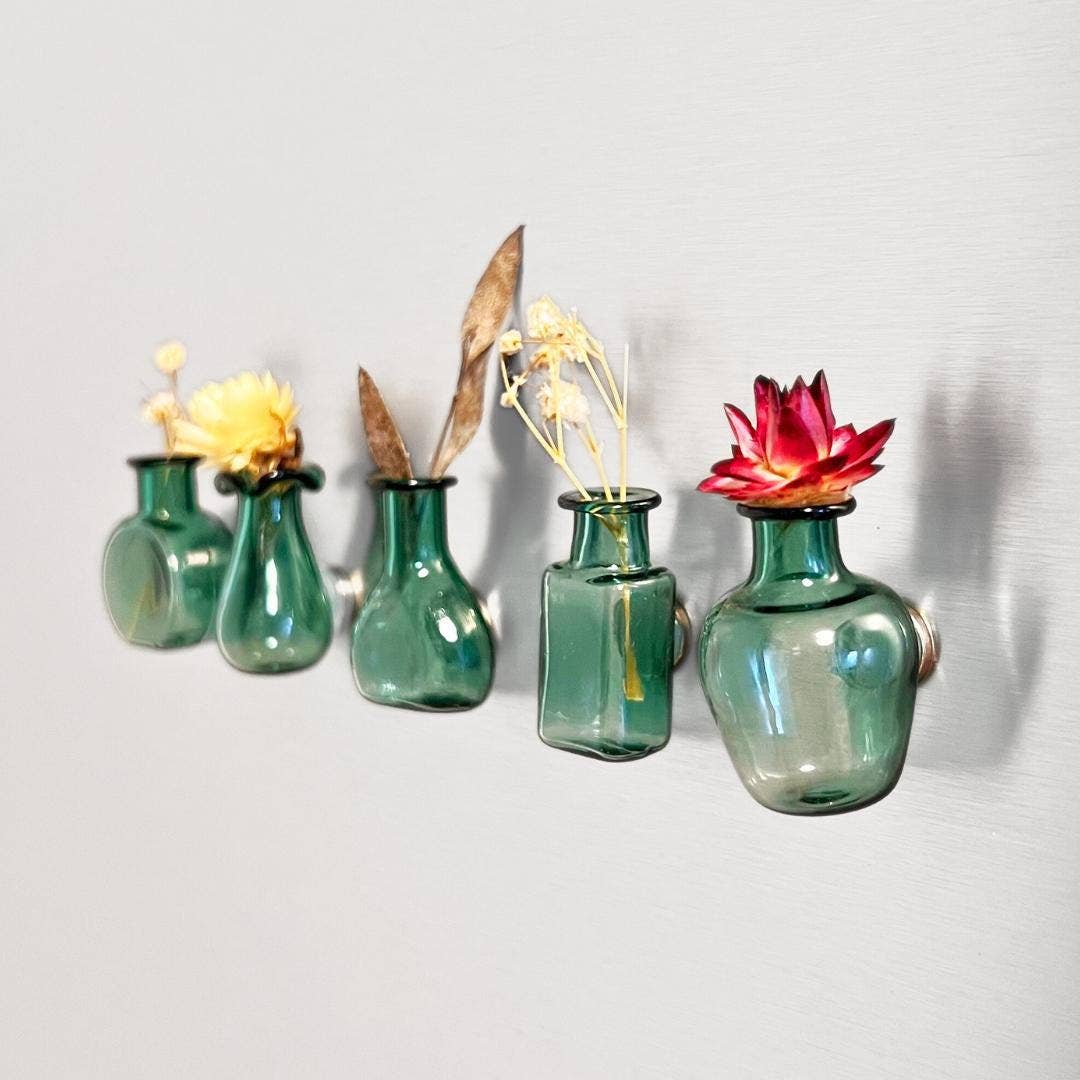 Five small translucent green glass vases mounted on a light gray wall, each holding dried flowers or stems in different shapes and colors.