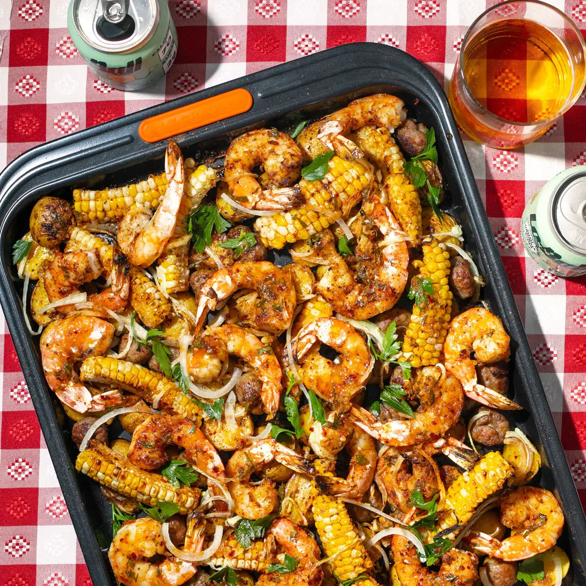 Tray of grilled shrimp and corn on a checkered tablecloth with drinks.