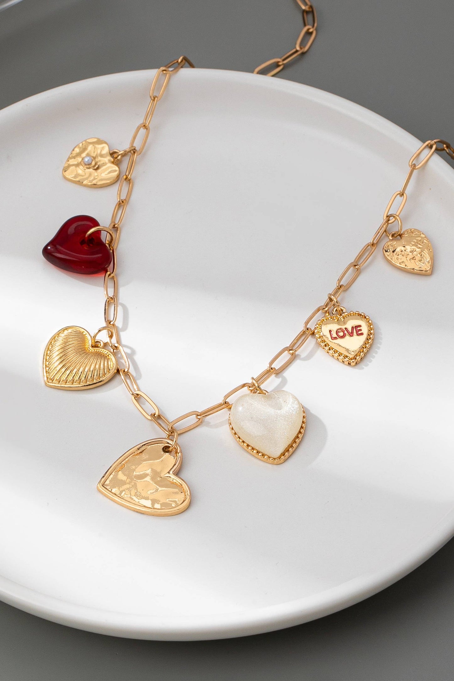 Gold chain link necklace with different heart shaped gold, red, and pearl charms, draped over a white plate