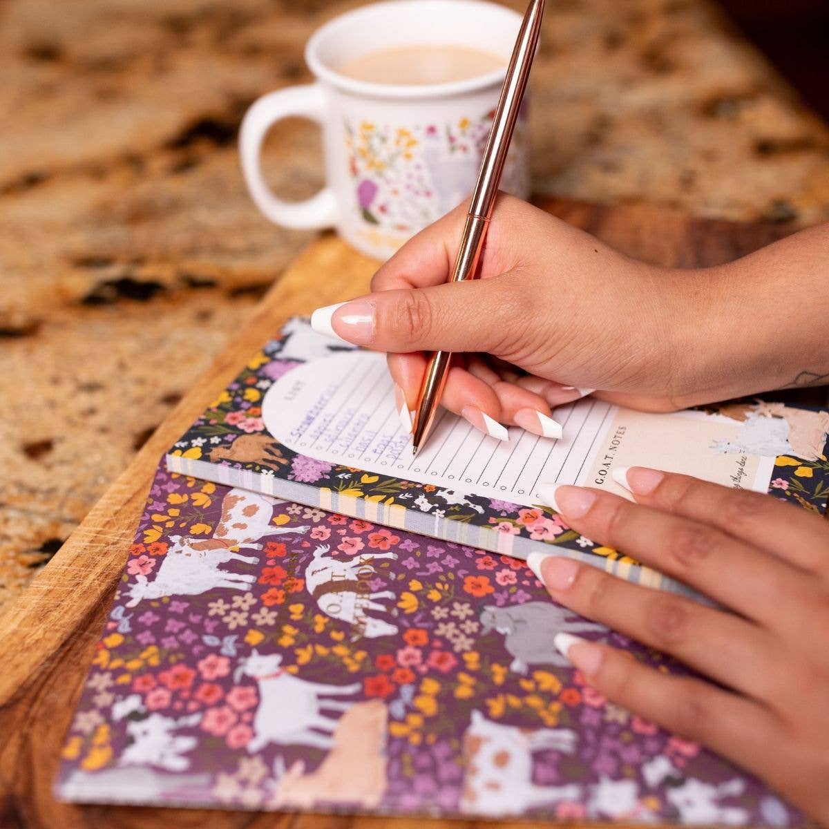 Person writing in a notebook with a floral cover on a wooden table with a cup of coffee.