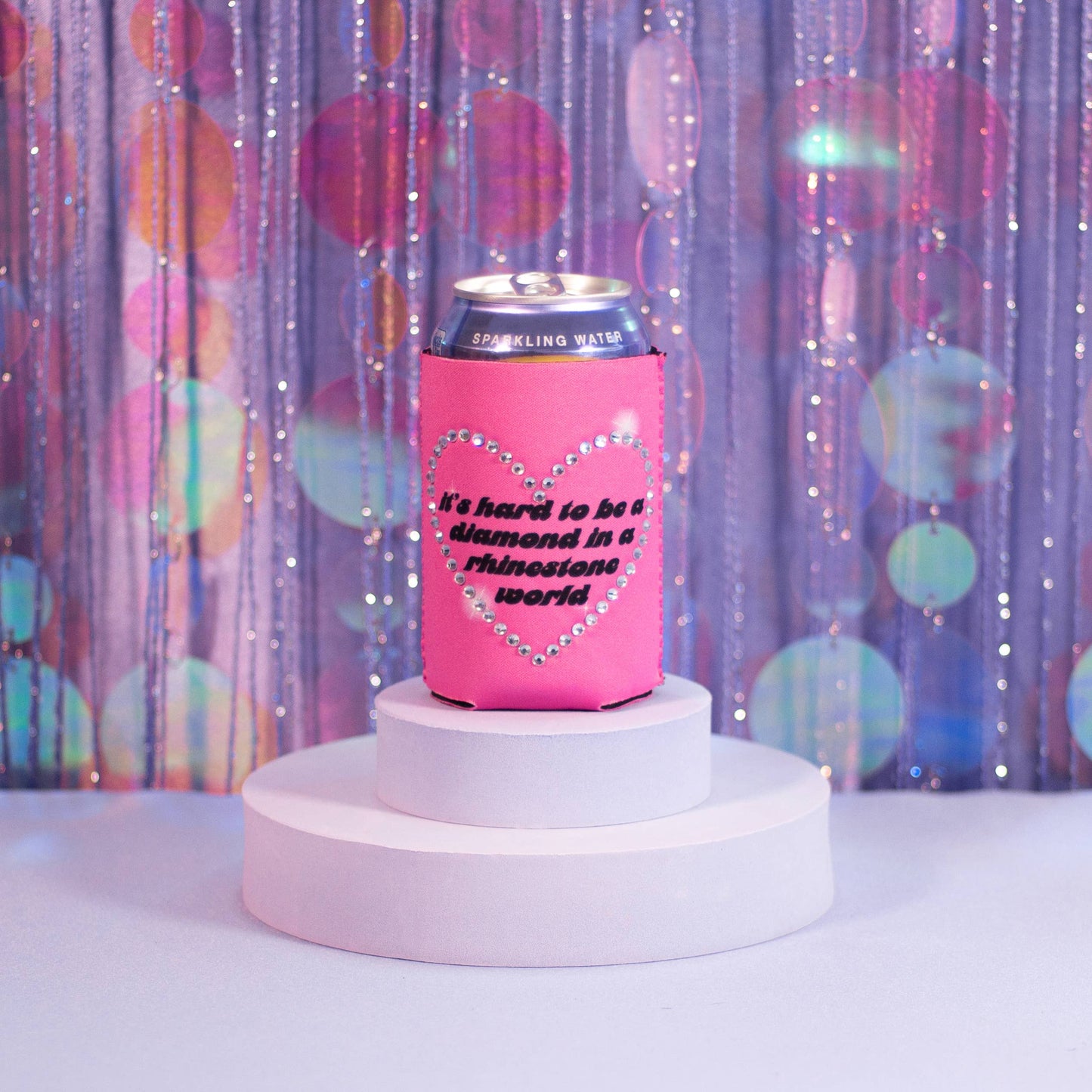 Pink can cooler with rhinestone heart design and text inside reading "It's Hard to be a diamond in a rhinestone world" on a white pedestal against a colorful curtain background