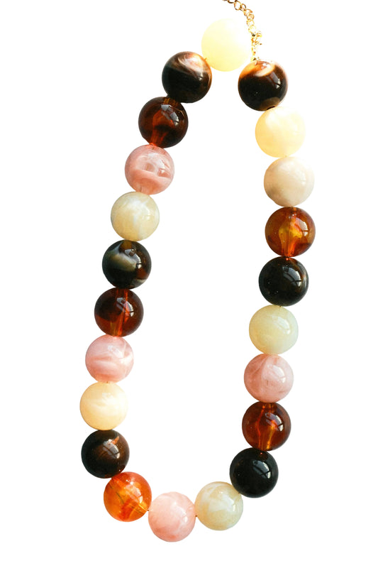A beaded necklace with alternating translucent amber-brown, glossy black, pale pink, cream, and orange round beads, displayed on a light blue-gray background with soft shadows.