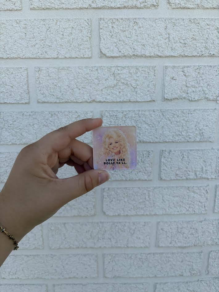 A hand holding a small square card featuring a portrait of Dolly Parton with the text 'LOVE LIKE DOLLY Y'ALL' against a white brick wall background.