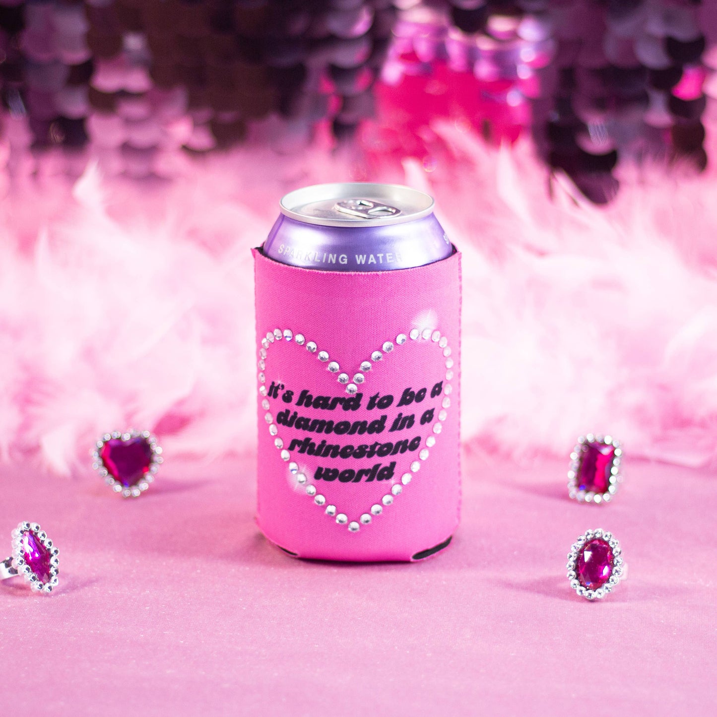 Pink can cooler with rhinestone heart design and text inside reading "It's Hard to be a diamond in a rhinestone world" surrounded by decorative pink hearts on a pink background.