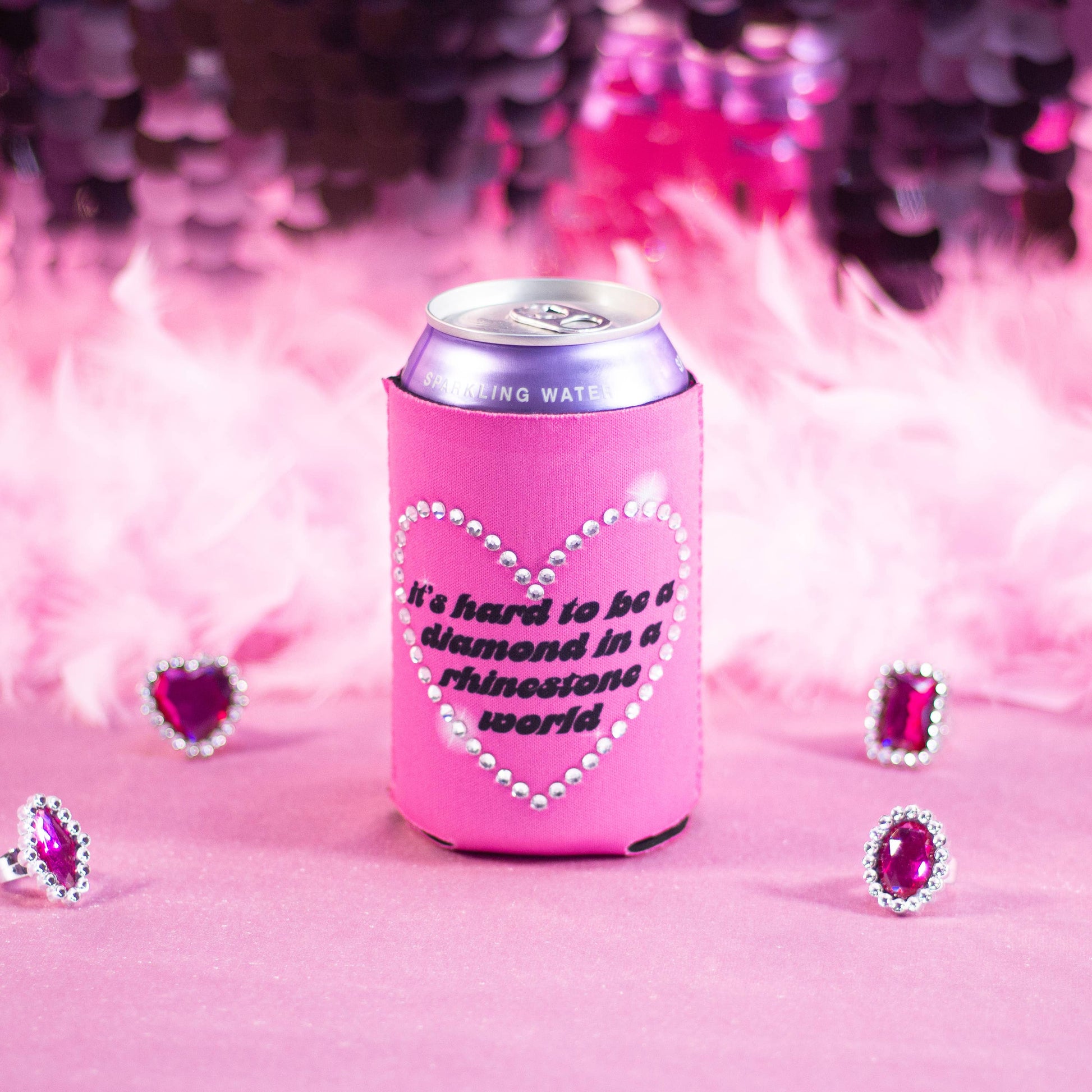 Pink can cooler with rhinestone heart design and text inside reading "It's Hard to be a diamond in a rhinestone world" surrounded by decorative pink hearts on a pink background.