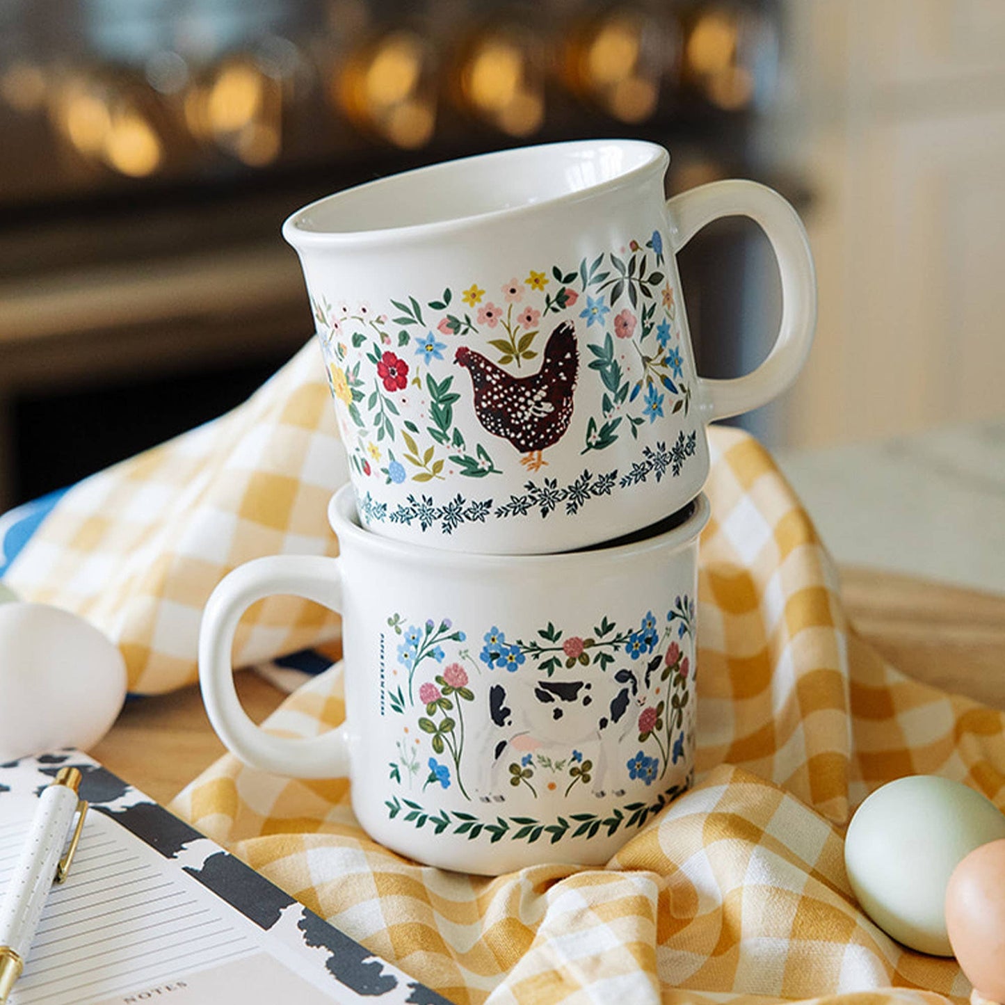 Two ceramic mugs with floral and animal designs stacked on a checkered tablecloth.