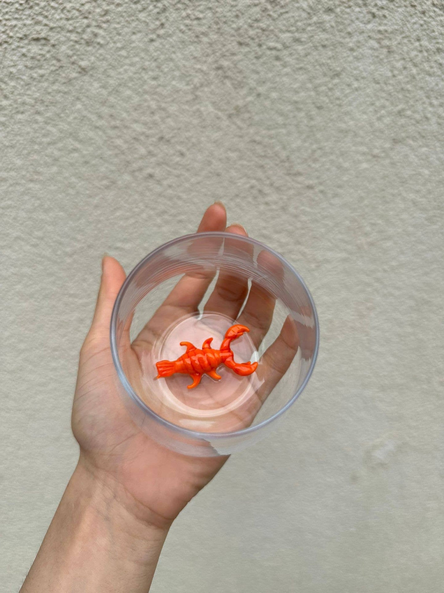 Hand holding a clear plastic cup with an orange toy inside against a plain background